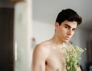 Vista laterale del giovane ragazzo senza maglietta con fiori bianchi freschi in mano guardando la fotocamera su sfondo sfocato — Foto stock