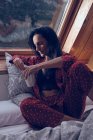 Attraktive Frau im Pyjama mit einem Becher Heißgetränk, während sie auf einem bequemen Bett in der Nähe des Fensters im gemütlichen Zimmer sitzt — Stockfoto