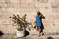 Atractiva joven hembra en gafas de sol de pie cerca de una antigua pared de piedra y una bonita planta en maceta en un día soleado - foto de stock