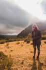 Joven dama de sombrero y chaqueta de esquí con mochila y bastón de pie en el campo cerca de la montaña en las nubes con sol en Isoba, Castilla y León, España - foto de stock