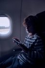 Чарівний хлопчик в повсякденному вбранні дивиться фільм на сучасному планшеті, сидячи біля вікна в темній кабіні сучасних літаків — стокове фото
