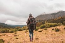 Vista posteriore della signora in cappello e giacca da sci con zaino e bastone da passeggio camminare sul prato vicino alla montagna in nuvole a Isoba, Castiglia e Leon, Spagna — Foto stock