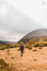 Вид сзади на даму в шляпе и лыжной куртке с рюкзаком и тростью ходьба по лугу возле горы в облаках в Исобе, Кастилии и Леоне, Испания — стоковое фото
