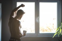 Вдумчивый человек, стоящий у окон с чашкой кофе — стоковое фото