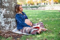 Усміхнена вагітна приваблива жінка сидить під деревом — стокове фото