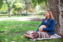 Усміхнена вагітна приваблива жінка сидить під деревом — стокове фото