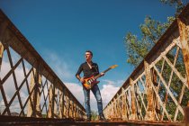 Ragazzo adulto con chitarra elettrica in piedi sul ponte intemperie e guardando altrove nella giornata di sole in campagna — Foto stock