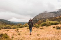 Back view of lady in hat and ski jacket with knapsack and walking stick walking on meadow near mountain in clouds in Isoba, Castile and Leon, Spain — Stock Photo