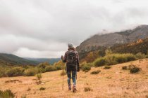 Vista trasera de la señora en sombrero y chaqueta de esquí con mochila y bastón caminando en prado cerca de la montaña en las nubes en Isoba, Castilla y León, España - foto de stock