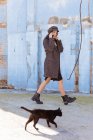 Stylish young woman in coat and cap talking on mobile phone while walking near blue brick wall and black cat — Stock Photo