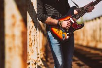 Colheita Adulto cara com guitarra elétrica de pé na ponte weathered no dia ensolarado no campo — Fotografia de Stock
