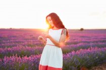 Lächelnde junge Frau zeigt Blumen zwischen violettem Lavendelfeld im Gegenlicht — Stockfoto
