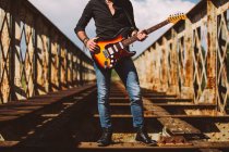 Culture Homme adulte avec guitare électrique debout sur le pont altérée par une journée ensoleillée à la campagne — Photo de stock
