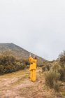 Vista lateral da jovem senhora em casaco de esqui amarelo com câmera usando telefone celular em mãos altas perto de colina e céu nublado em Isoba, Castela e Leão, Espanha — Fotografia de Stock