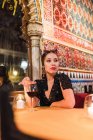 Attraktive junge Frau in Kleid sitzt am Tisch neben Glas mit Getränken und brennenden Kerzen in luxuriösen Raum des Cafés durch Mosaik dekoriert — Stockfoto