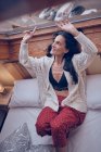 Sensual female in knitted jacket and bra smiling and pointing at window glass while sitting on comfortable bed at home — Stock Photo