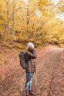 Vista lateral de uma senhora de chapéu e casaco de esqui com mochila e bengala segurando câmera ao nível do rosto no caminho entre a floresta de outono em Isoba, Castela e Leão, Espanha — Fotografia de Stock