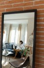 Reflexão da jovem mulher sentada no sofá com lençol perto da mesa baixa com pincéis e desenhar sobre tela na sala de luz — Fotografia de Stock