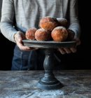 Crop human holding tray with set of fresh cakes with powdered sugar on wooden table in darkness — Stock Photo