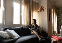 Молодая привлекательная женщина в платье сидит на диване в квартире и смотрит в окно — стоковое фото