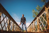 Adulte guy avec guitare électrique debout sur weathered pont et regarder loin par jour ensoleillé dans campagne — Photo de stock