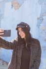 Charming Hispanic lady taking selfie with mobile phone in front of shabby wall — Stock Photo