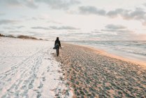Back view of lady in coat and hat with backpack going on sand shore near snow and water in Klaipeda, Lithuania — Foto stock