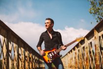 Adult guy with electric guitar standing on weathered bridge and looking away on sunny day in countryside — Stock Photo