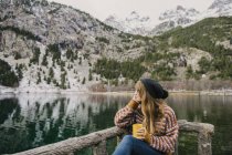 Jeune femme assise sur un banc et regardant loin près d'une vue imprenable sur la surface de l'eau entre de hautes montagnes avec des arbres dans la neige dans les Pyrénées — Photo de stock