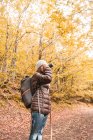 Бічний вид на жінку в капелюсі і гірськолижній куртці з рюкзаком і ходячою палицею, яка тримає камеру на рівні обличчя на пішохідному шляху між осіннім лісом в Ісобі, Кастилія і Леон, Іспанія. — стокове фото