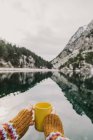 Рука человека держит желтую кружку рядом с удивительным видом на водную поверхность между высокими горами с деревьями в снегу и облачным небом в Пиренеях — стоковое фото