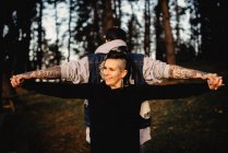 Молодой человек в татуировках, стоящий спиной к спине и держащийся за руки с улыбающейся женщиной в парке на размытом фоне — стоковое фото