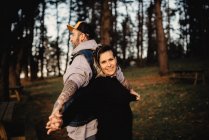 Young man in tattoos with standing back to back and holding hands with smiling woman in park on blurred background — Stock Photo