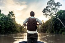 Back view young guy with knapsack standing on bow of ship sailing on river between exotic forest on shores in Malaysia — Stock Photo