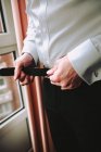 Side view of crop hands of male buttoning leather belt on black trousers near window — Stock Photo