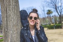 Vista lateral de una joven mujer hipster riendo de pie y apoyada en un árbol en el parque en un día soleado mientras usa un teléfono móvil - foto de stock