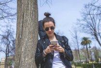 Side view of a young laughing hipster woman standing and leaning on a tree at park in sunny day while using a mobile phone — Stock Photo
