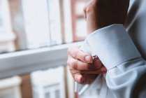 Side view of crop hands of male buttoning up white shirt sleeve near window — Stock Photo