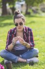 Vista frontale di una giovane hipster seduta sull'erba in un parco mentre regge un fiore in una giornata di sole — Foto stock