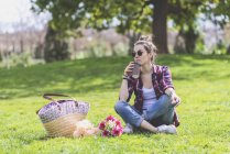 Вид спереди на молодую хипстершу, сидящую на траве и смотрящую в парк, держа кружку на вынос в солнечный день — стоковое фото
