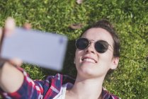 Sopra la vista di una giovane donna hipster sorridente sdraiata sull'erba in una giornata di sole in un parco mentre si fa un selfie con un telefono cellulare — Foto stock