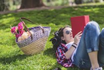 Vista lateral de una mujer feliz hipster acostado en la hierba en el día soleado en el parque mientras lee un libro rojo - foto de stock