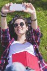 Vista superior de una joven mujer hipster sonriente tumbada en la hierba en un día soleado en un parque mientras toma una selfie con un teléfono móvil - foto de stock