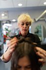 Stilista anziano rendendo acconciatura attraente giovane signora nel salone di parrucchiere — Foto stock