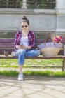 Front view of a young hipster woman sitting on a park bench relaxing in a sunny day while using a mobile phone — Stock Photo