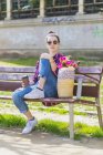 Vorderansicht einer jungen Hipster-Frau, die an einem sonnigen Tag auf einer Parkbank sitzt und in die Kamera blickt — Stockfoto