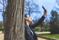Вид сбоку женщины, стоящей и опирающейся на дерево в парке в солнечный день, когда она использует мобильный телефон, чтобы сделать селфи — стоковое фото