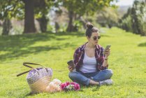 Передній вигляд молодої жінки-хіпстера в сонцезахисних окулярах, сидячи на траві в парку, використовуючи мобільний телефон — стокове фото