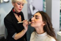Stylist rouging Lippen durch Pinsel zu attraktiven Frau mit Make-up auf verschwommenem Hintergrund — Stockfoto