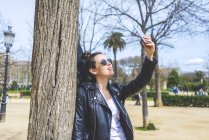 Вид сбоку женщины, стоящей и опирающейся на дерево в парке в солнечный день, когда она использует мобильный телефон, чтобы сделать селфи — стоковое фото